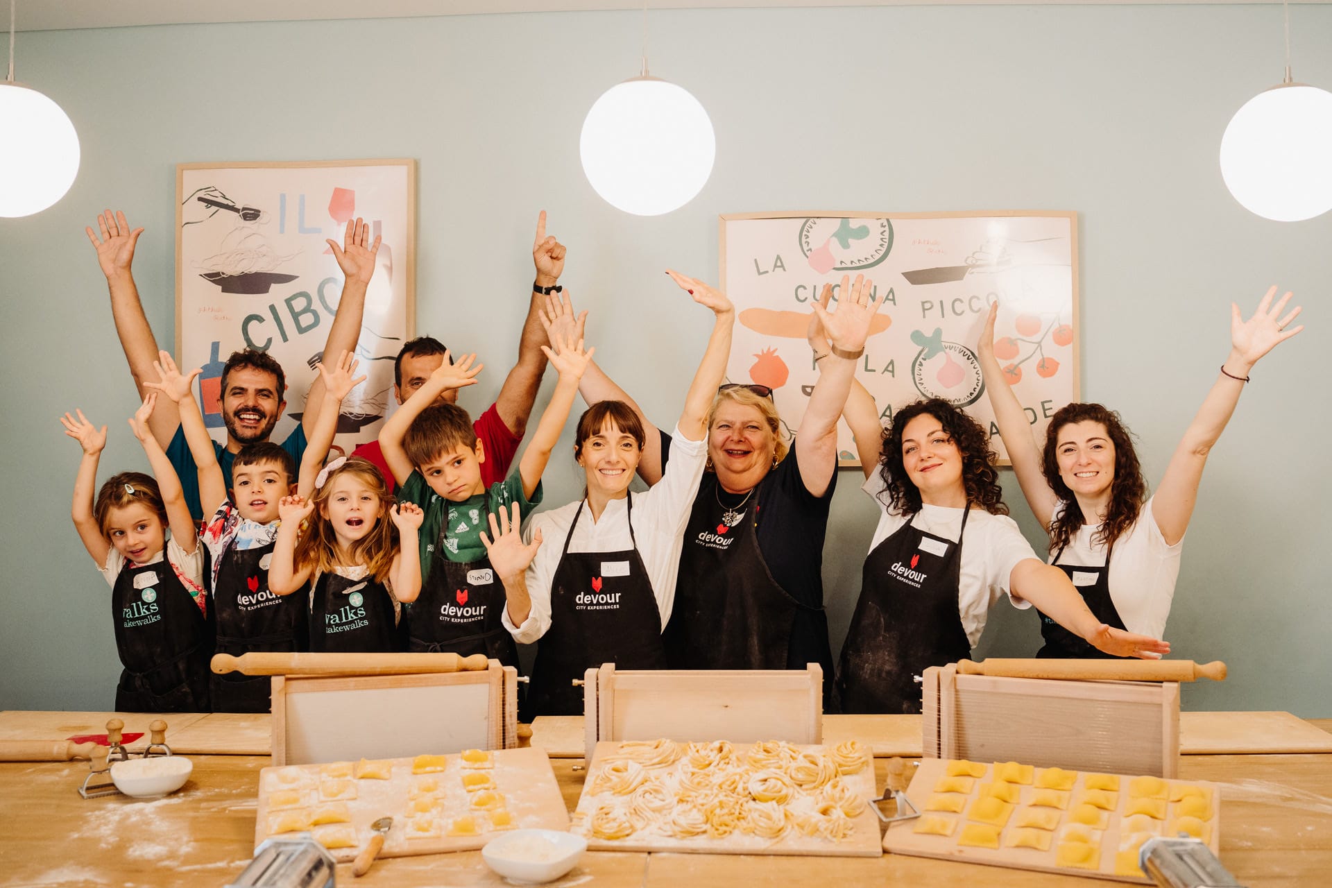 A joyful group of cooking class attendees, both kids and adults, celebrating around a table filled with handmade pasta shapes.